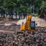Żółta koparka przygotowuje teren budowy domu w otoczeniu drzew