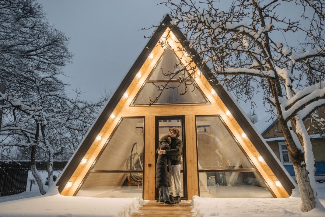 Para przed oświetlonym domem A-frame wśród ośnieżonych drzew Para przed oświetlonym domem A-frame wśród ośnieżonych drzew